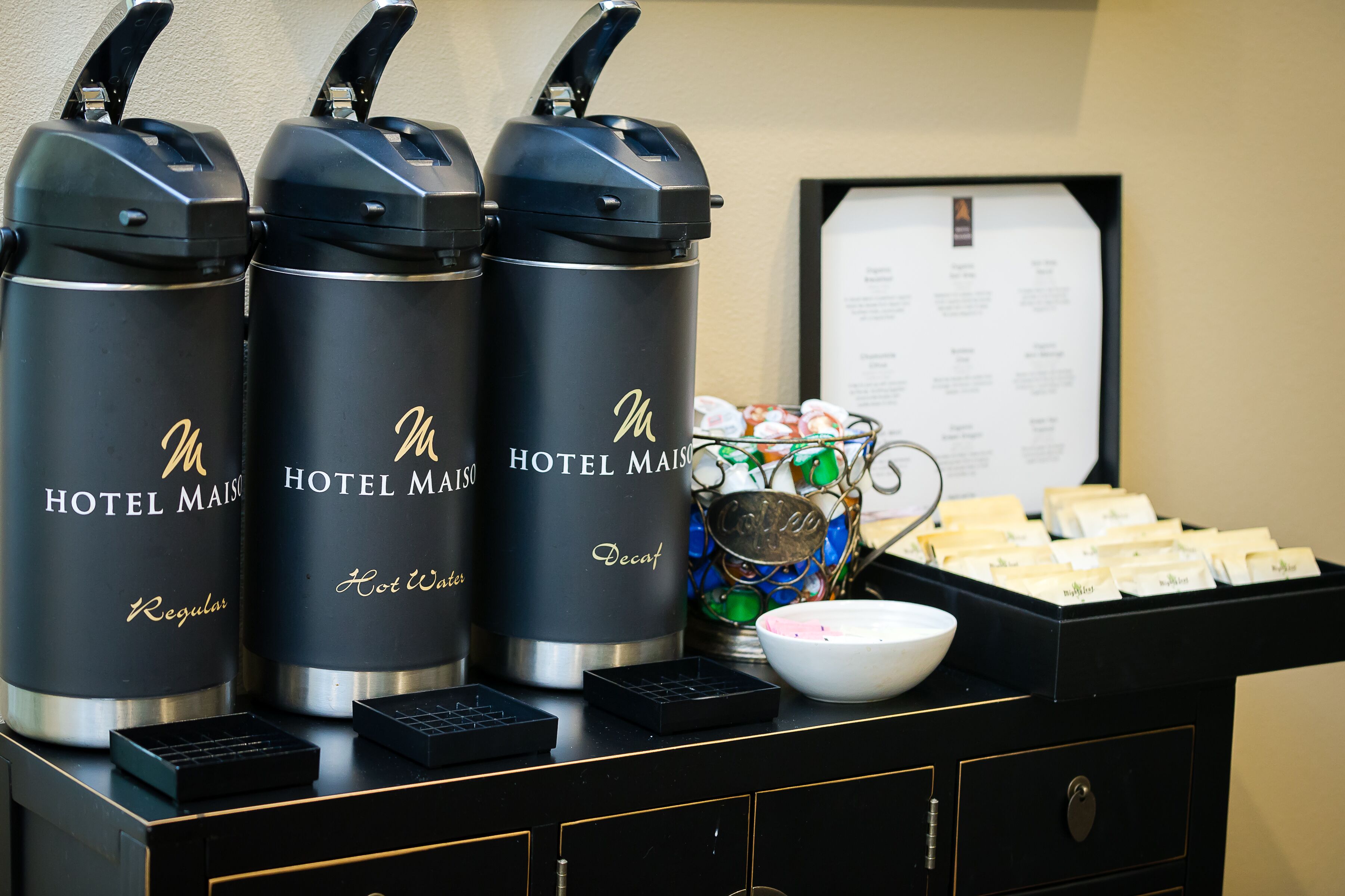 Tres dispensadores de agua negros de hotel con la etiqueta "HOTEL MAISON Descafeinado", un cuenco de aperitivos y un tablero de menú.