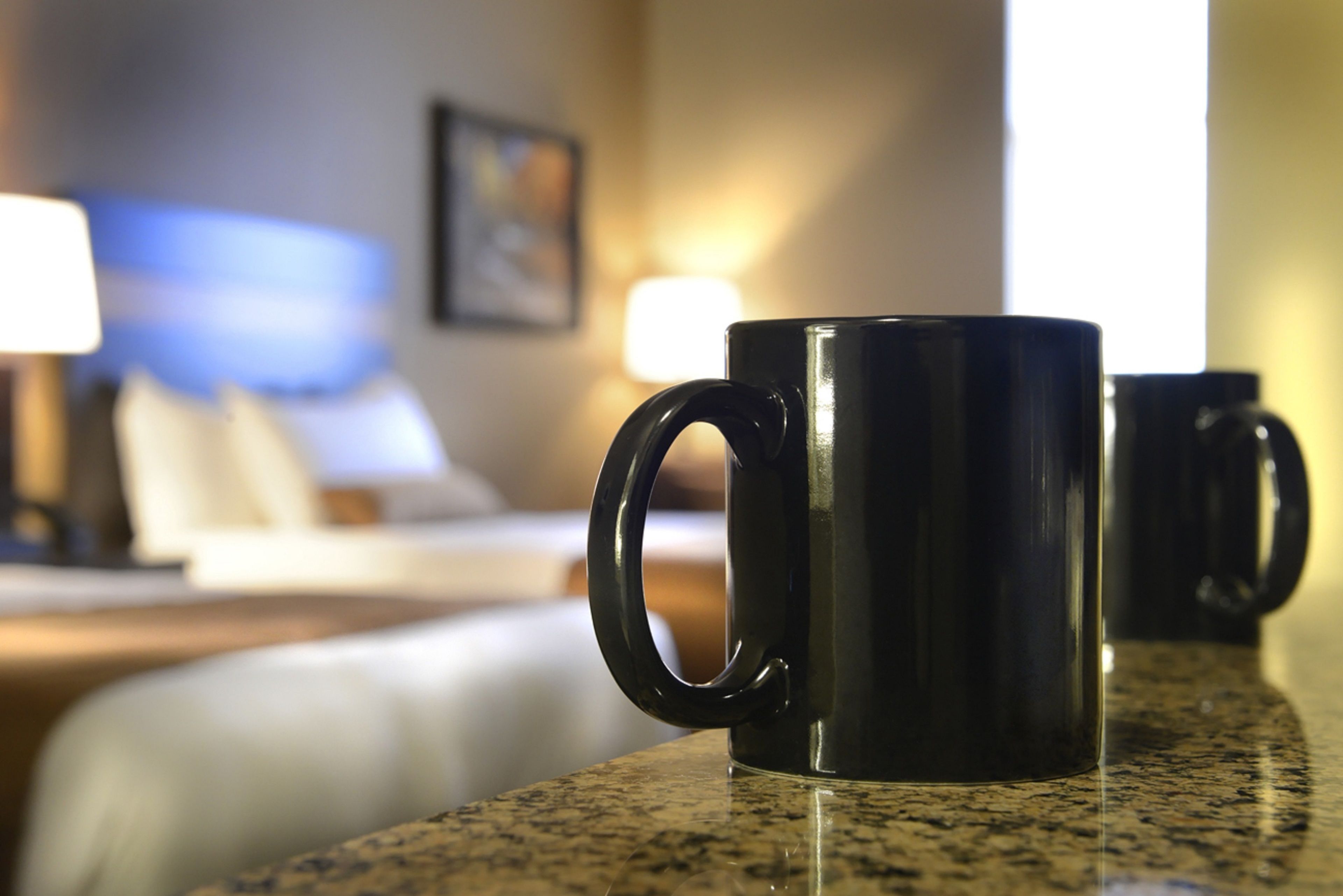 A black mug on a granite countertop in a hotel room with a bed and a nightstand in the background.