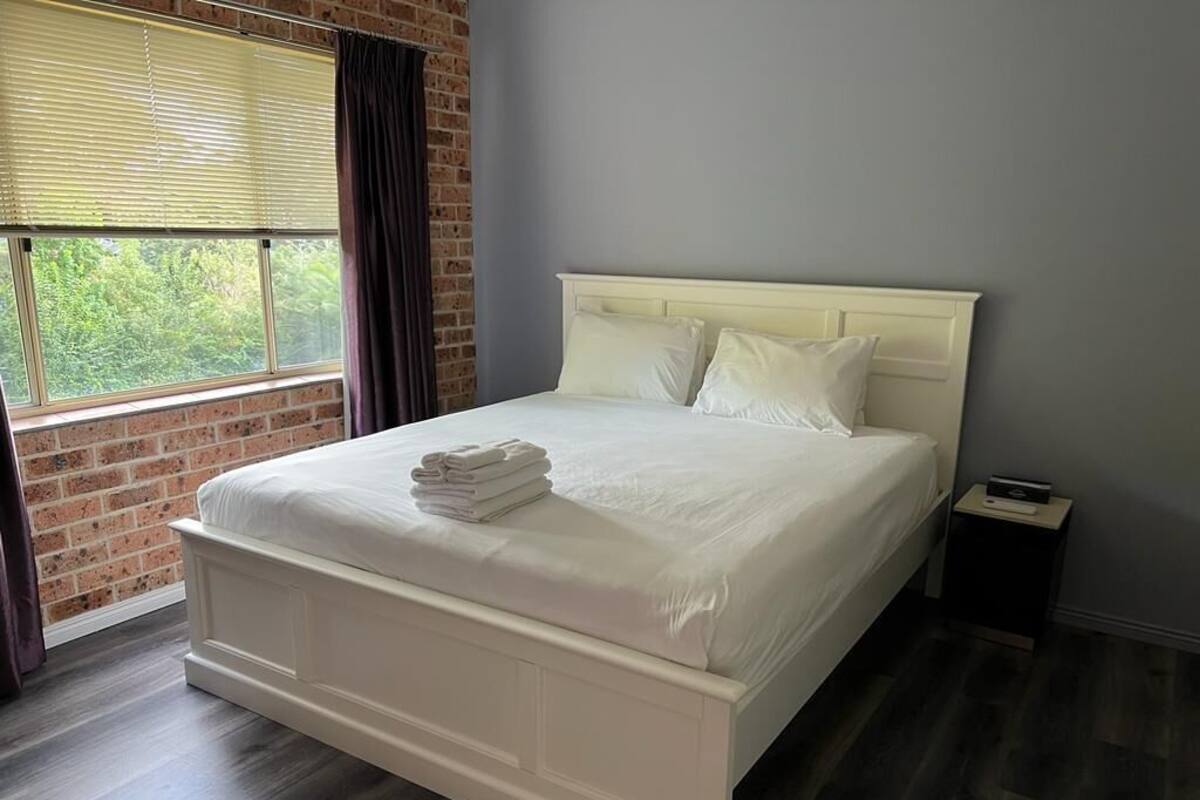 A neatly made bed with white linens and pillows, a small bedside table with a lamp, and a window with blinds.