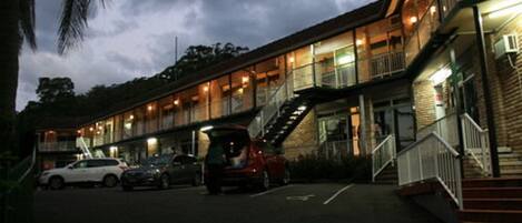 A multi-story motel with a central staircase, parking lot, and a row of cars parked in front.