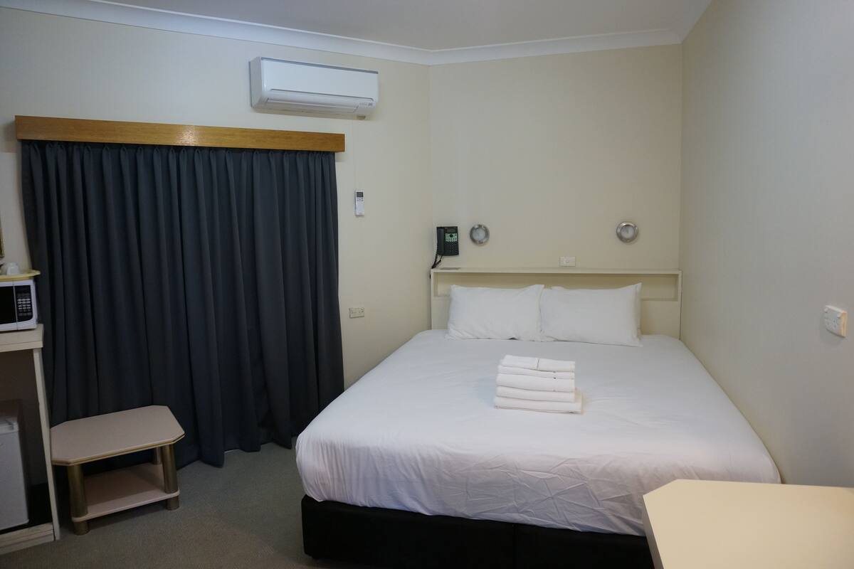 A hotel room with a bed, a bedside table, a desk, a chair, a microwave, and a curtain.