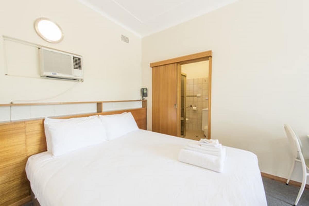 A hotel room with a bed, a wooden headboard, a wall-mounted air conditioner, a door leading to a bathroom, and a white chair.