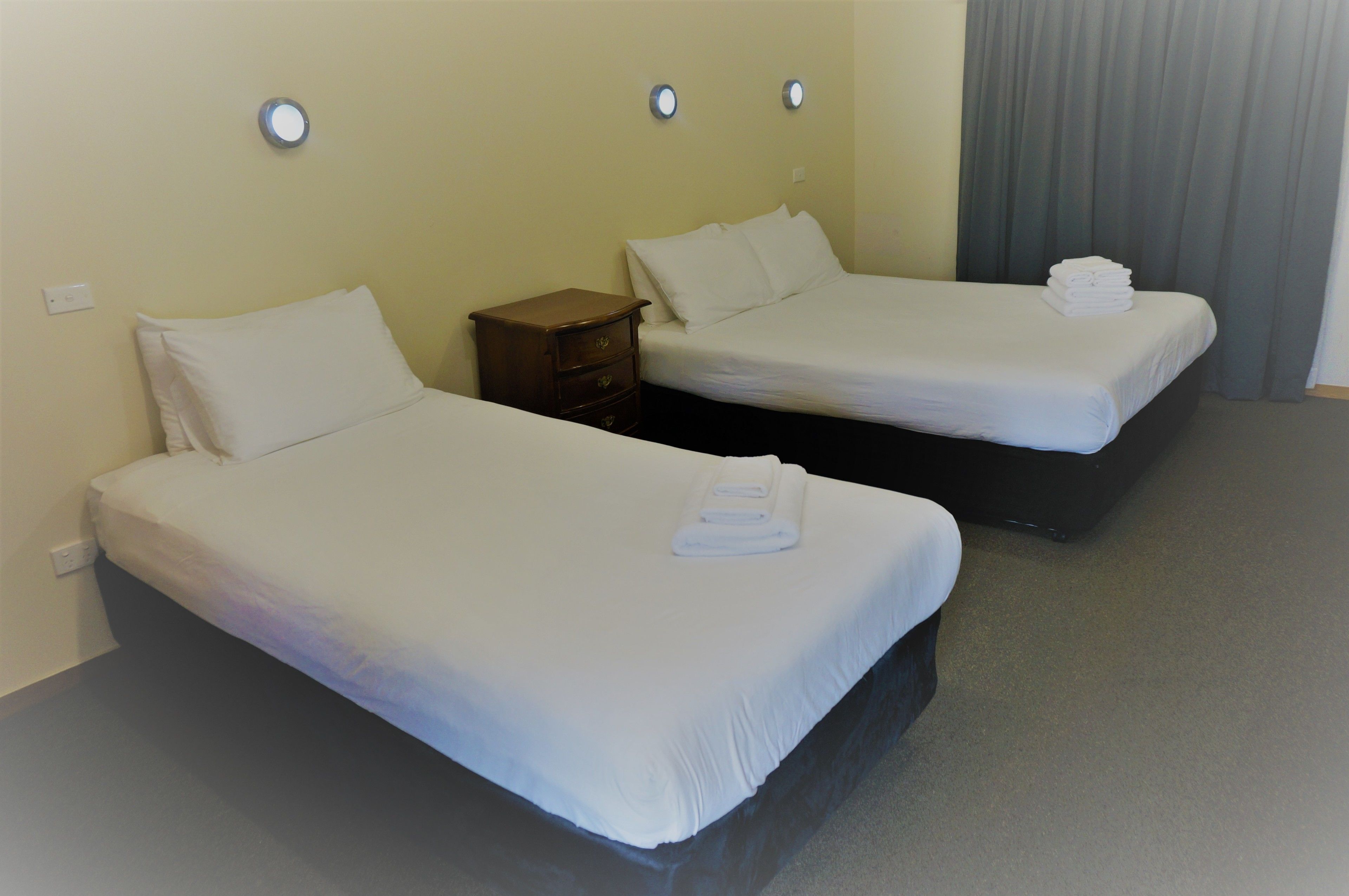 Two single beds in a room with a small wooden nightstand between them, a white wall, and a grey curtain.