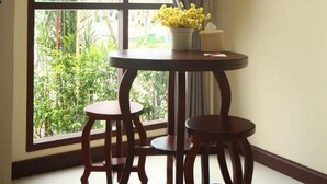 Une pièce avec une grande fenêtre, une table ronde en bois et deux chaises, ainsi qu’une plante en pot ornée de fleurs jaunes.