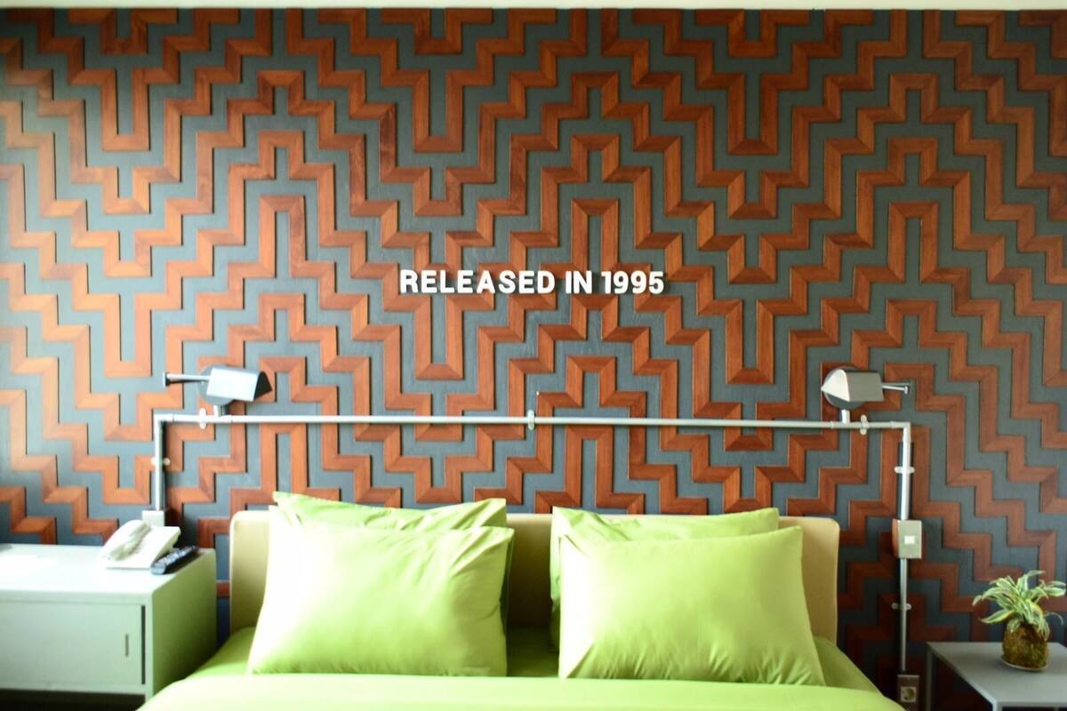 Habitación de hotel con pared de diseño geométrico, cama con almohadas verdes, mesita de noche con lámpara y una planta pequeña en el tocador.