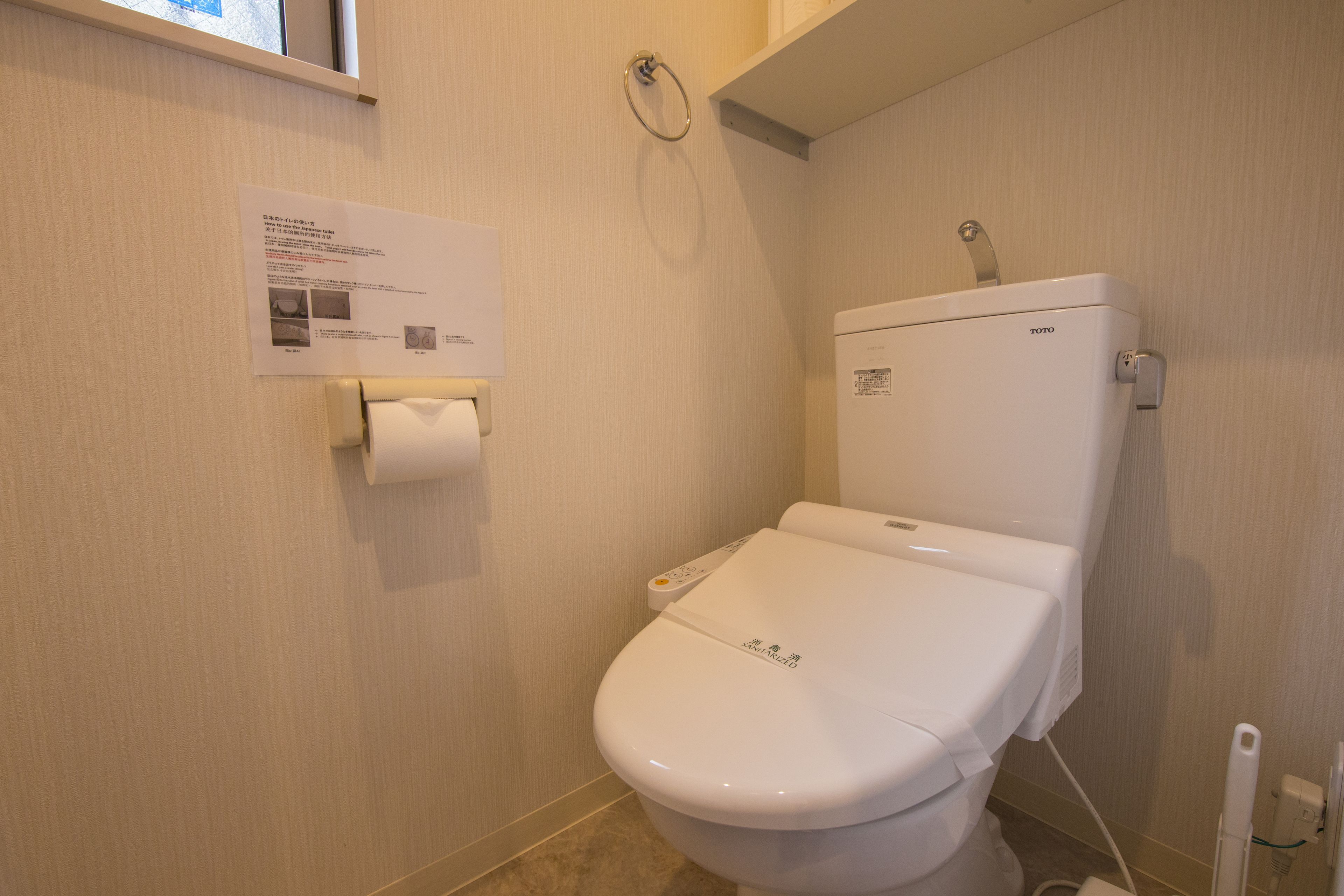 A standard white toilet with a TOTO brand label, a paper towel holder, and a hand sanitizer dispenser.