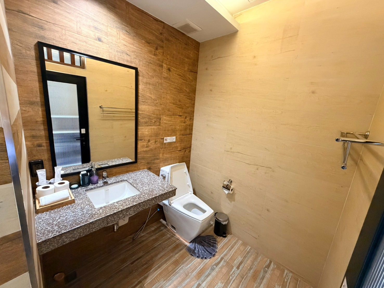A modern bathroom with a wooden wall, a large mirror, a sink, a toilet, and a shower area.