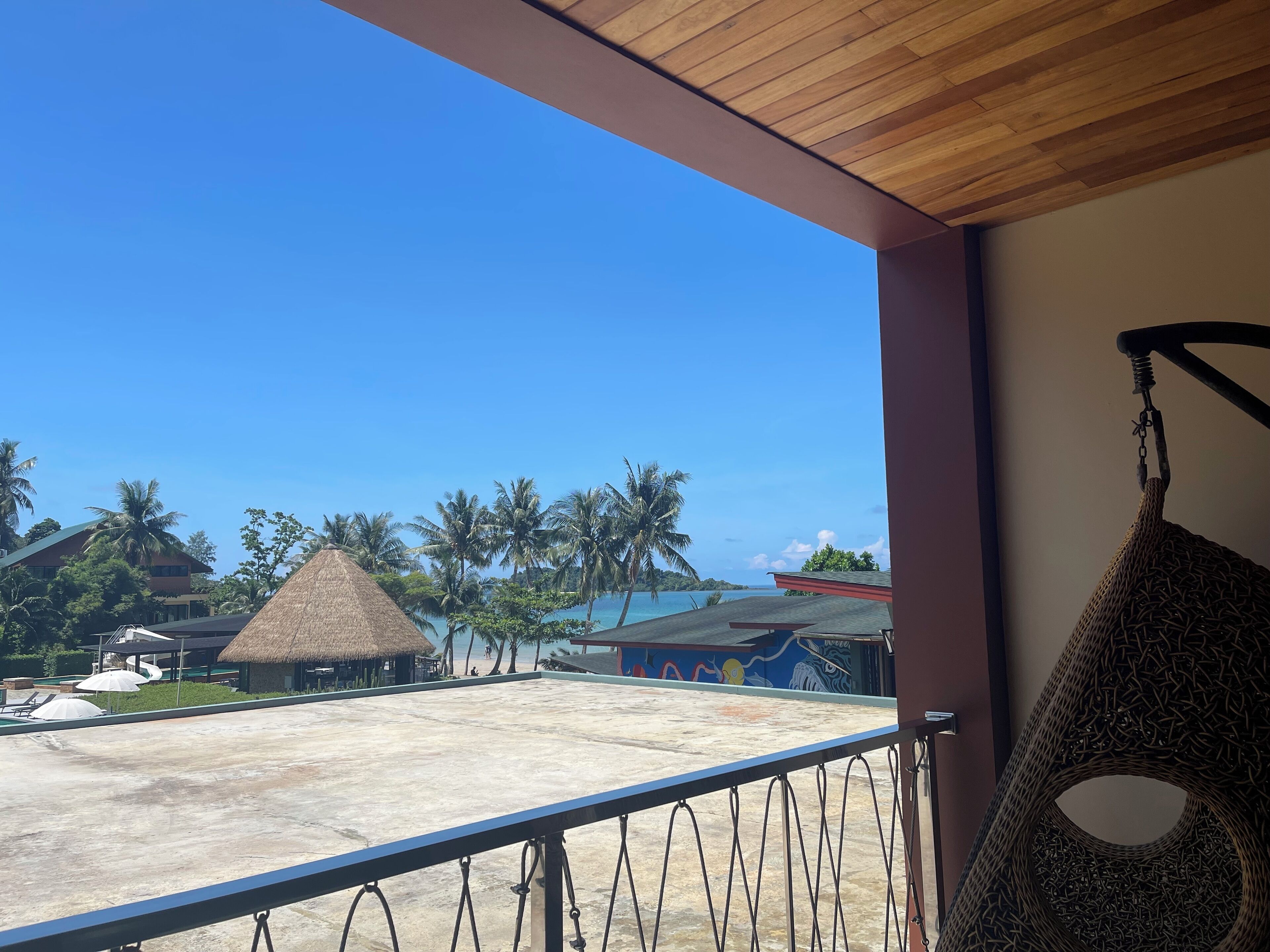 A balcony with a view of a beach, palm trees, and a thatched-roof building.