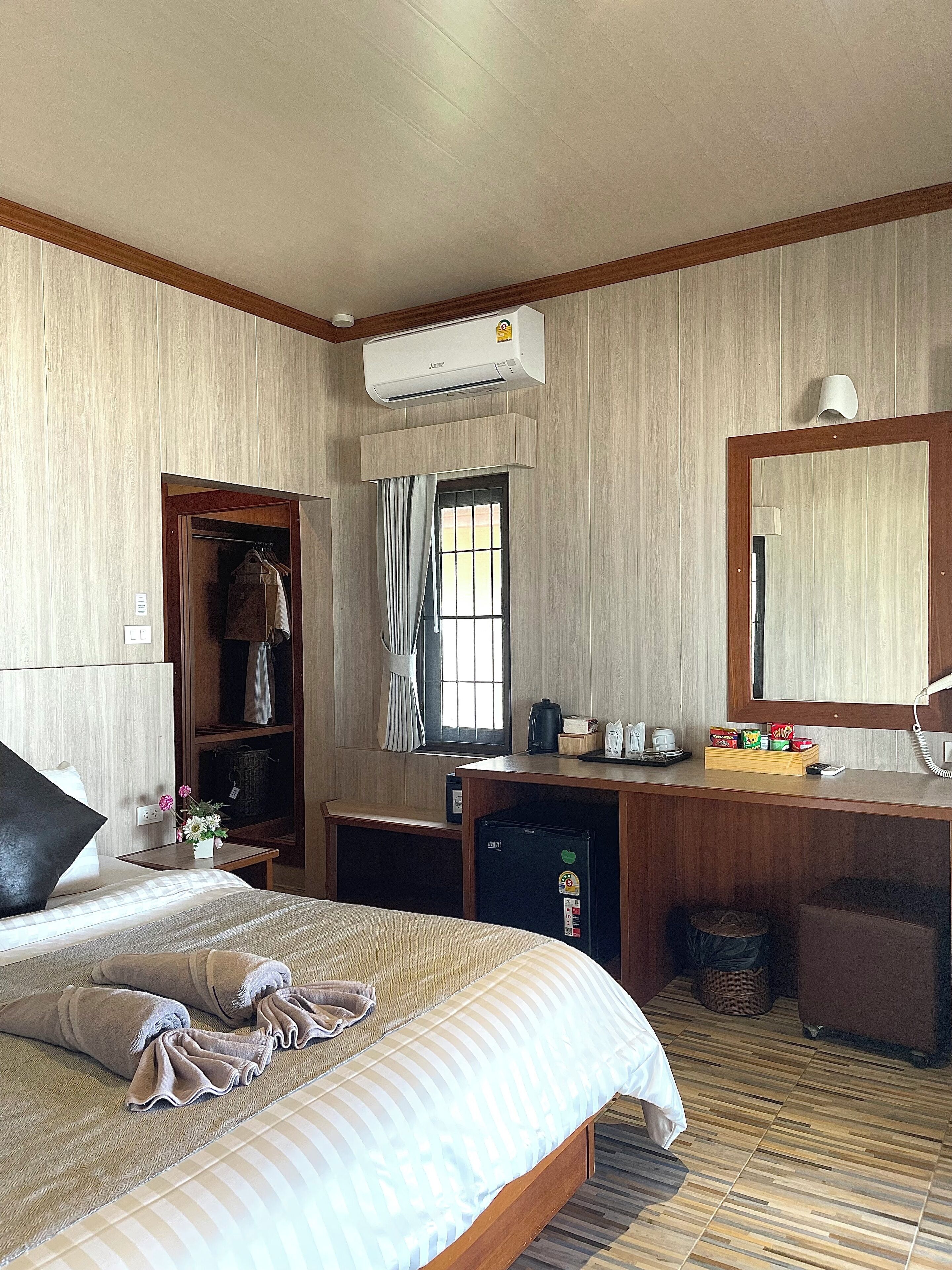 A hotel room with a bed, a desk, a wardrobe, an air conditioning unit, a mirror, and a window with curtains.