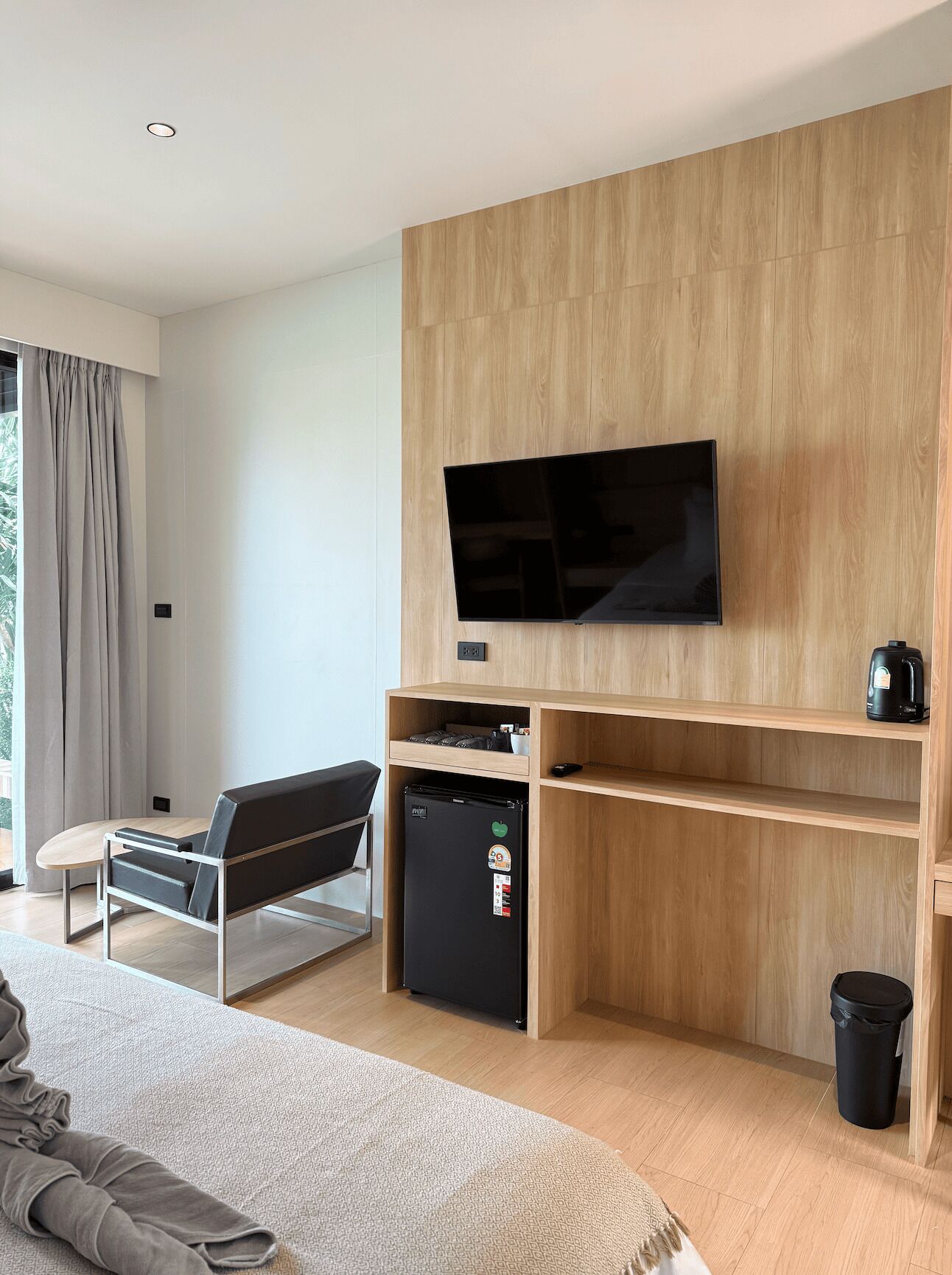 A modern living room with a flat-screen TV mounted on a wooden wall, a black refrigerator, a sofa, and a coffee machine on a shelf.