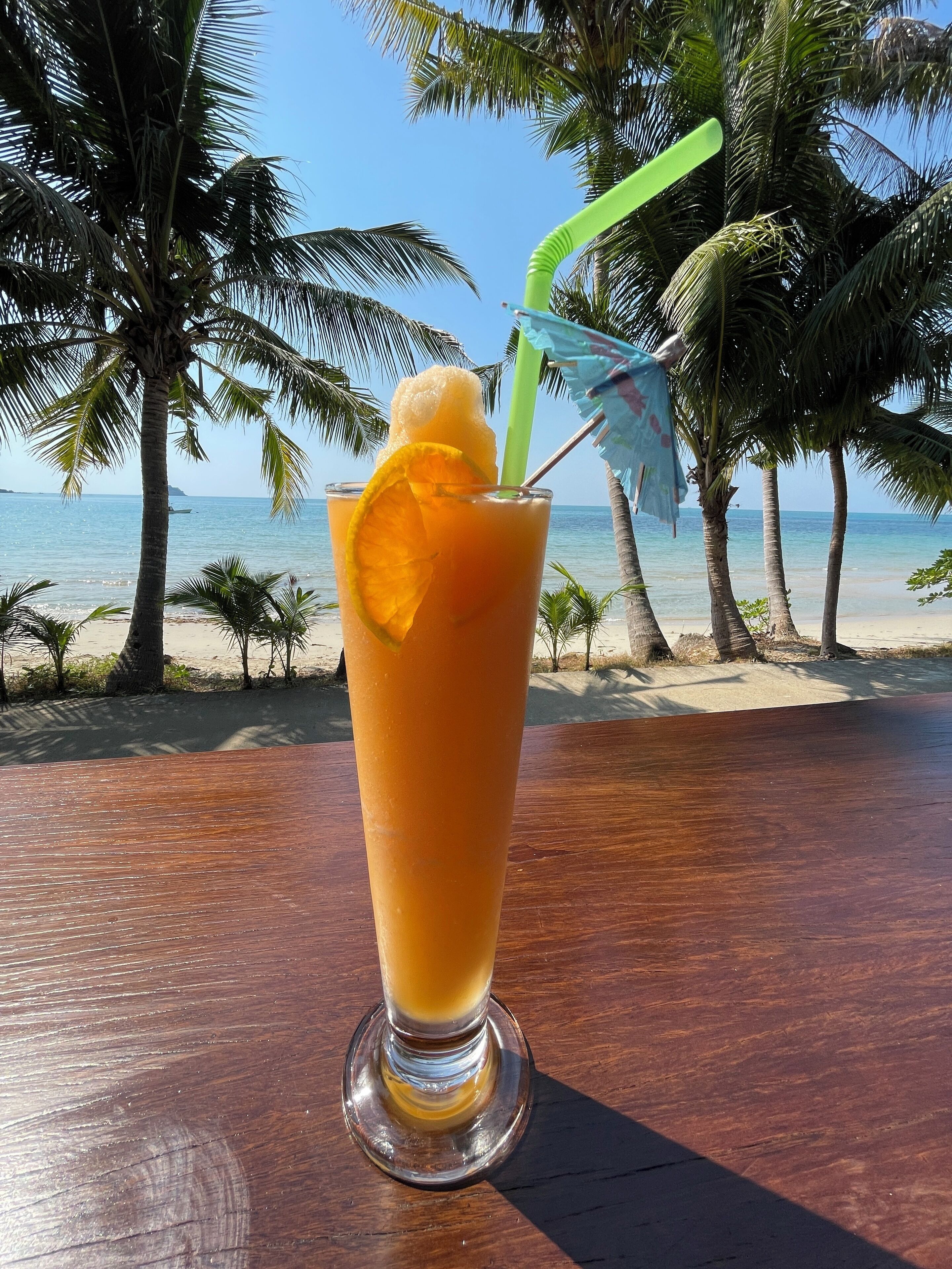 A tall glass of orange juice with a slice of orange and a straw on a wooden table with a beach view in the background.