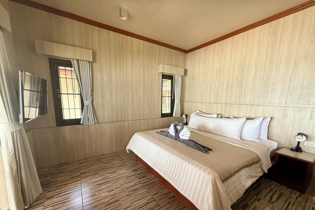 A hotel room with a large bed, two windows with curtains, a wooden floor, and a nightstand with a lamp.