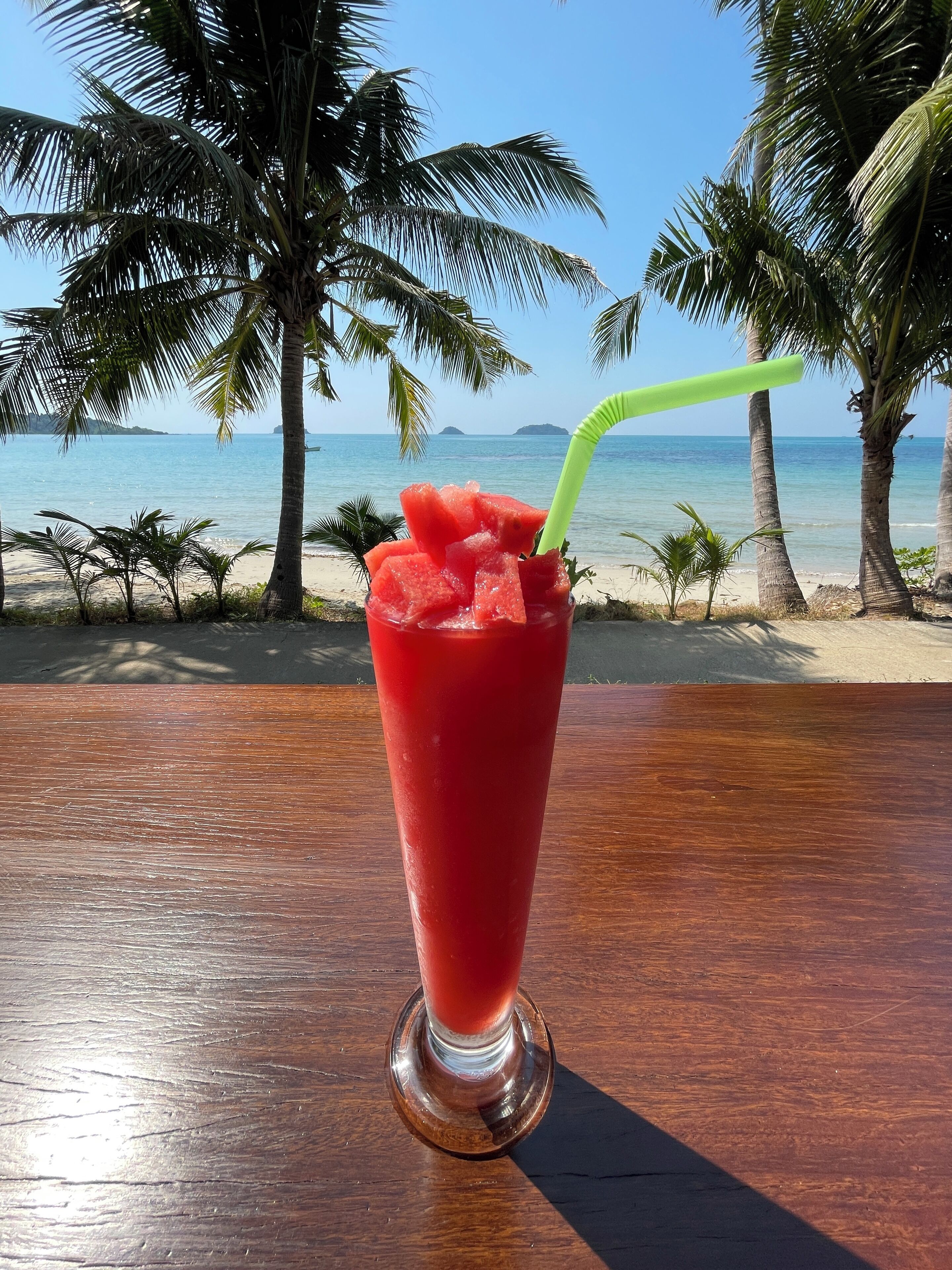 Un verre d’une boisson rouge contenant des morceaux de pastèque et une paille verte, posé sur une table en bois près d’une plage bordée de palmiers, avec l’océan en arrière-plan.