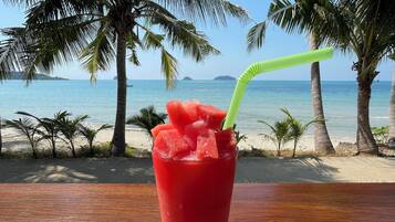 Un verre d’une boisson rouge contenant des morceaux de pastèque et une paille verte, posé sur une table en bois près d’une plage bordée de palmiers, avec l’océan en arrière-plan.