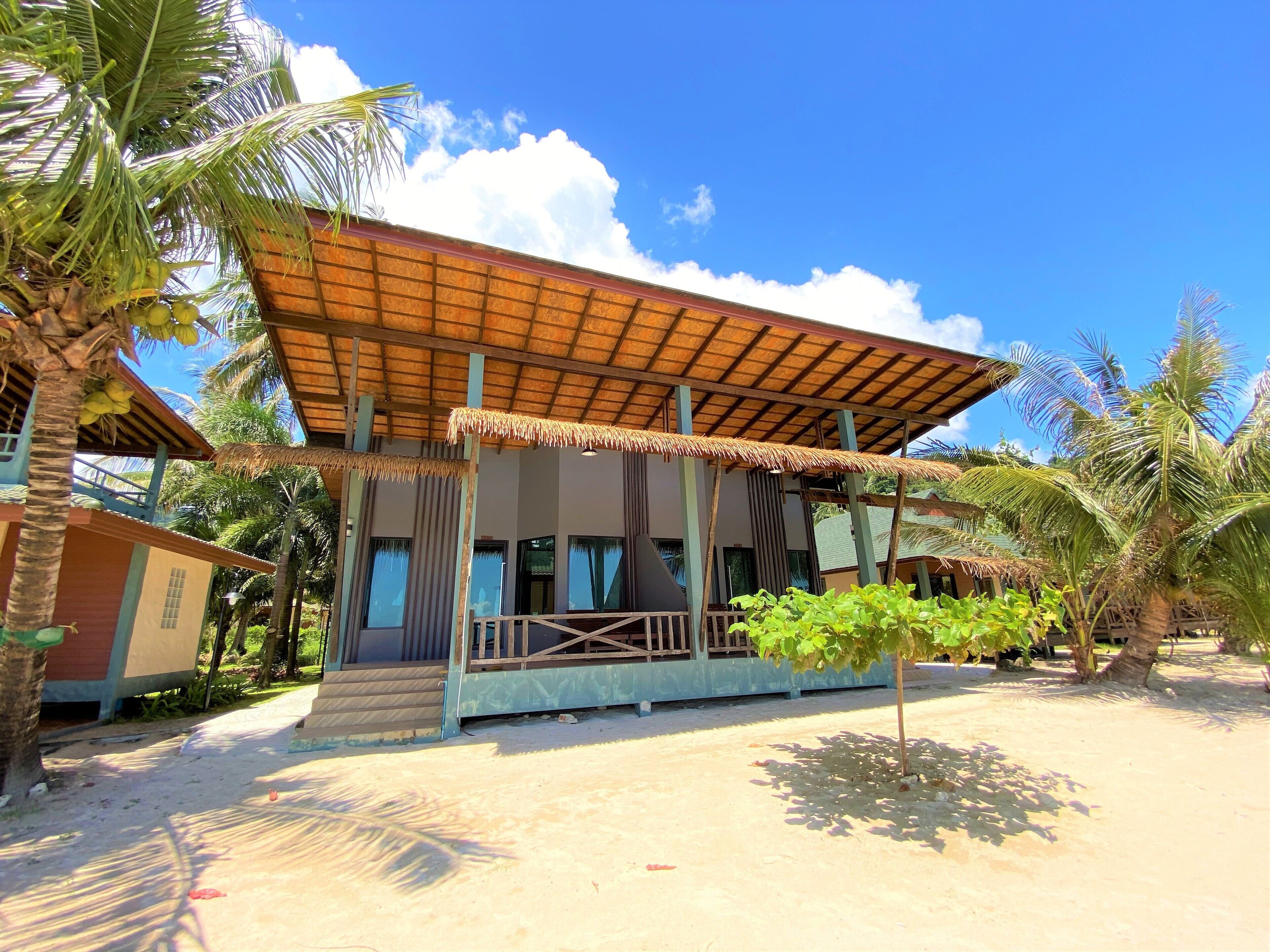 A two-story building with a thatched roof, surrounded by palm trees and a sandy ground.