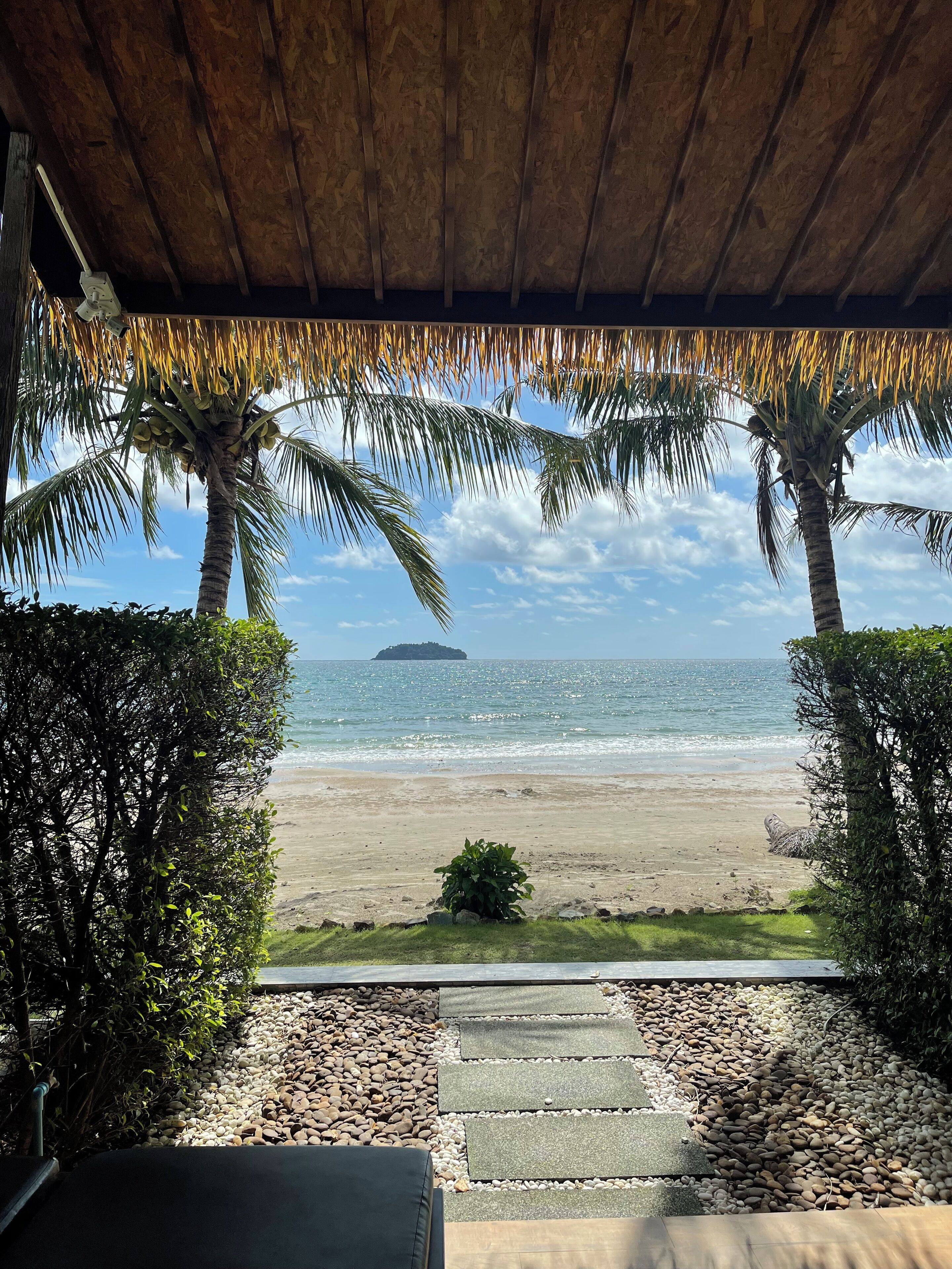 A covered outdoor area with a view of the beach and ocean, featuring palm trees and a distant island.