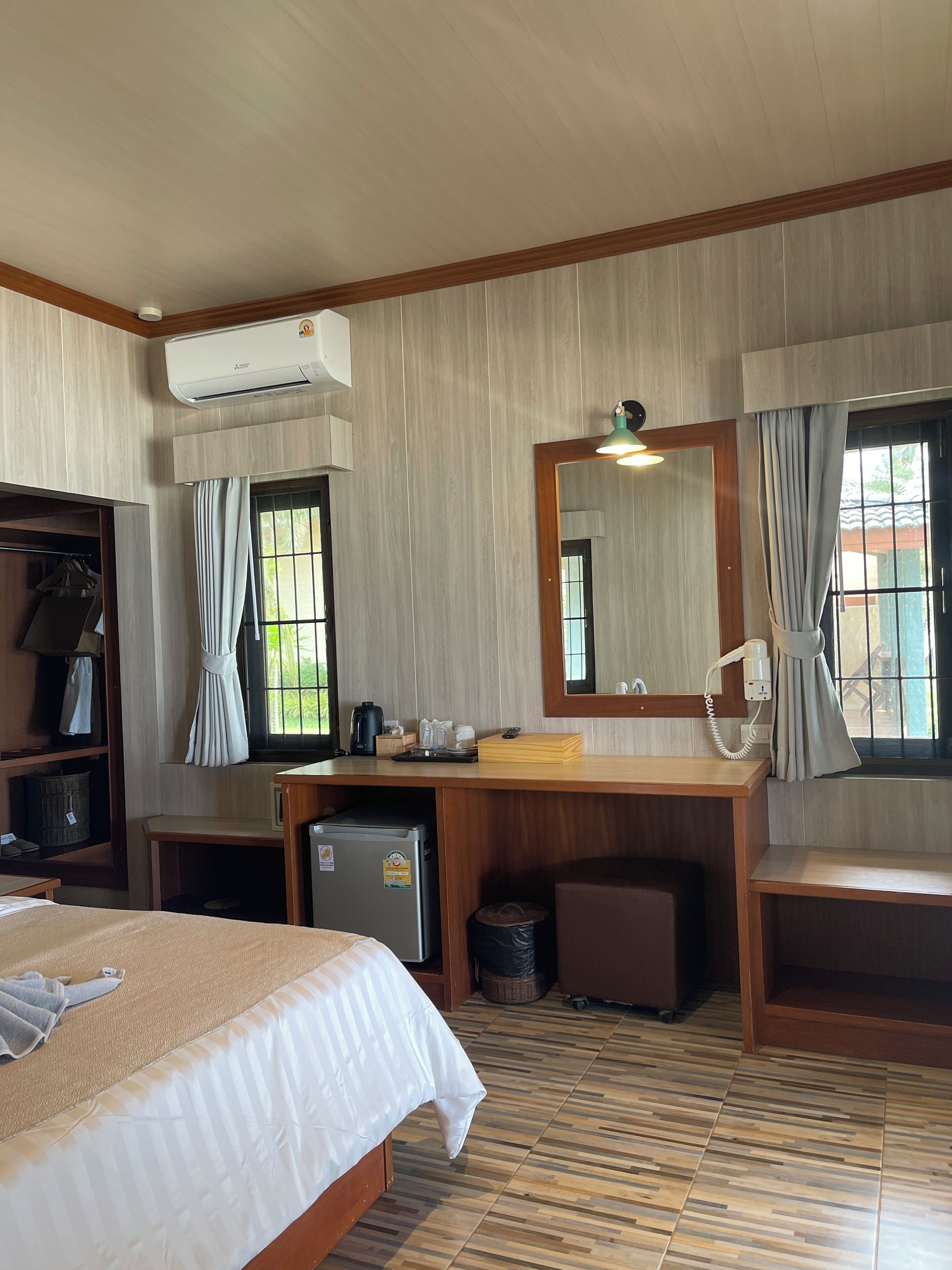 A hotel room with a bed, desk, and wardrobe. There is an air conditioning unit above the desk and a mirror above the desk.