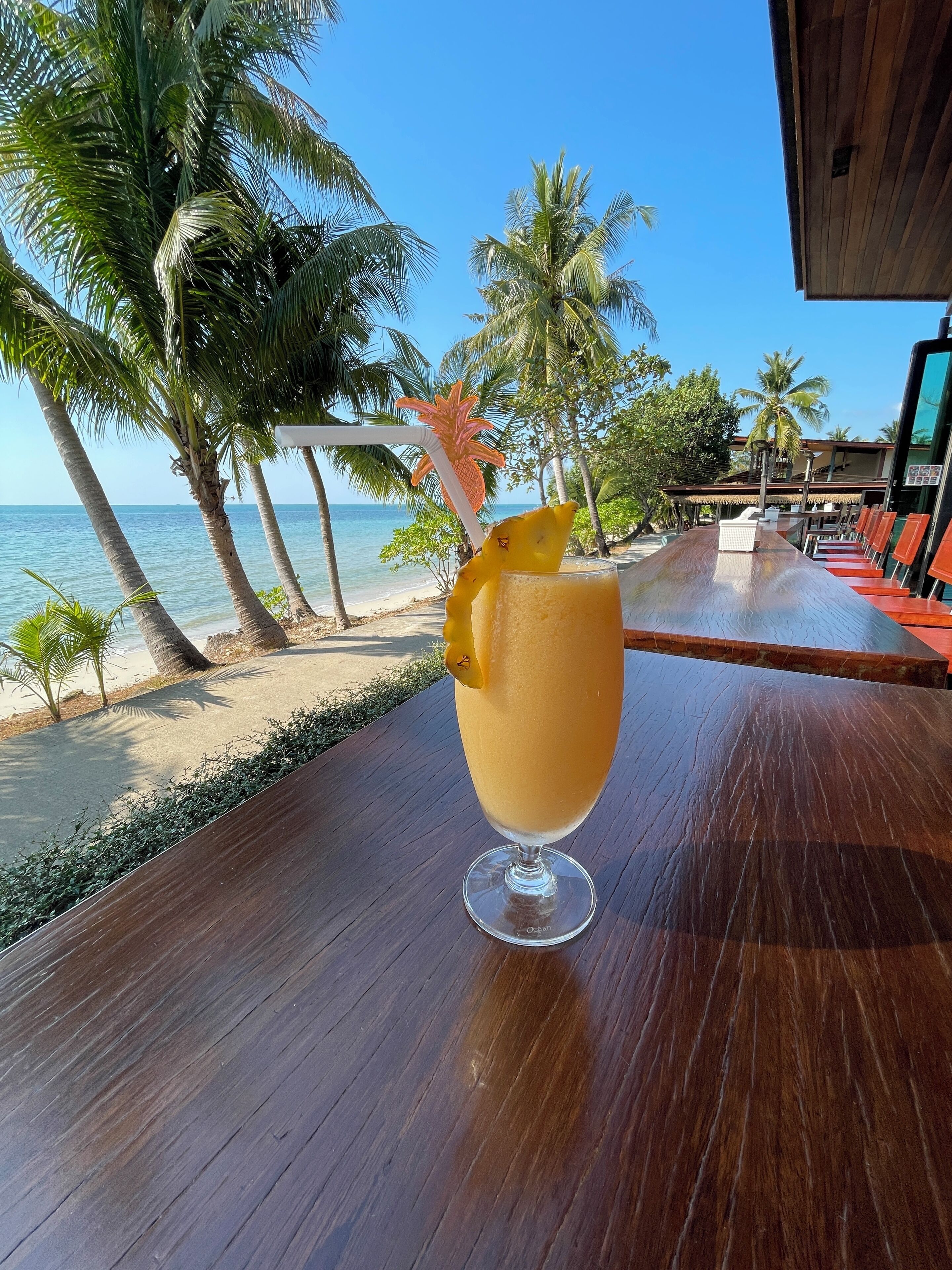 A tropical drink on a wooden table with a view of the beach and palm trees.
