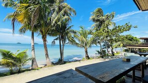 A beachside patio with wooden tables and benches, overlooking a calm sea and palm trees. - Kaibae Beach Resort (Ko Chang)