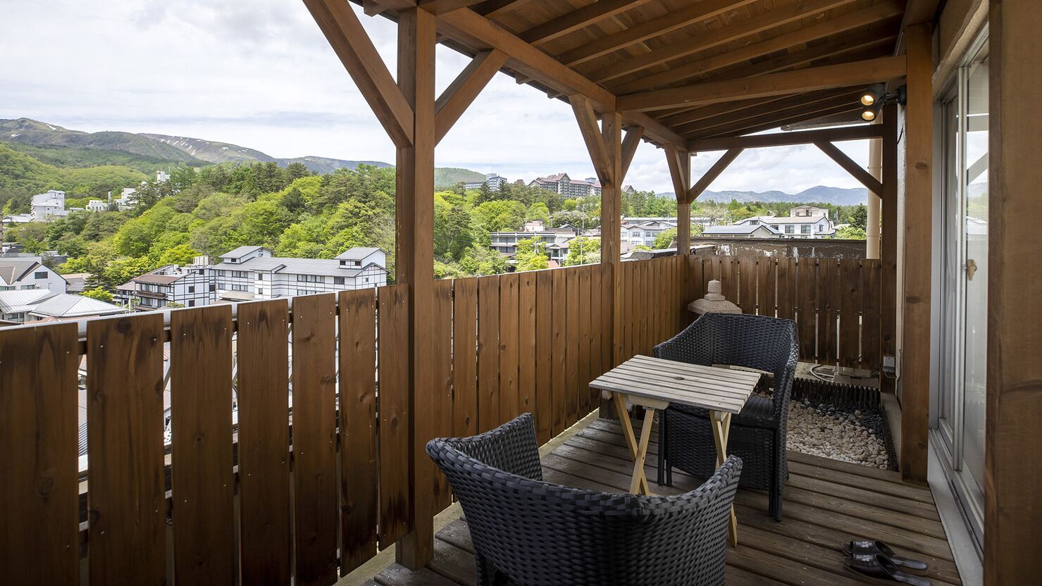 Um deck de madeira com móveis de vime e uma mesa, com vista para uma área residencial com casas e vegetação.