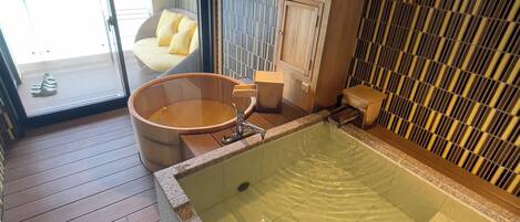 A modern bathroom with a large bathtub, a wooden floor, a glass door leading outside, and a seating area with cushions.