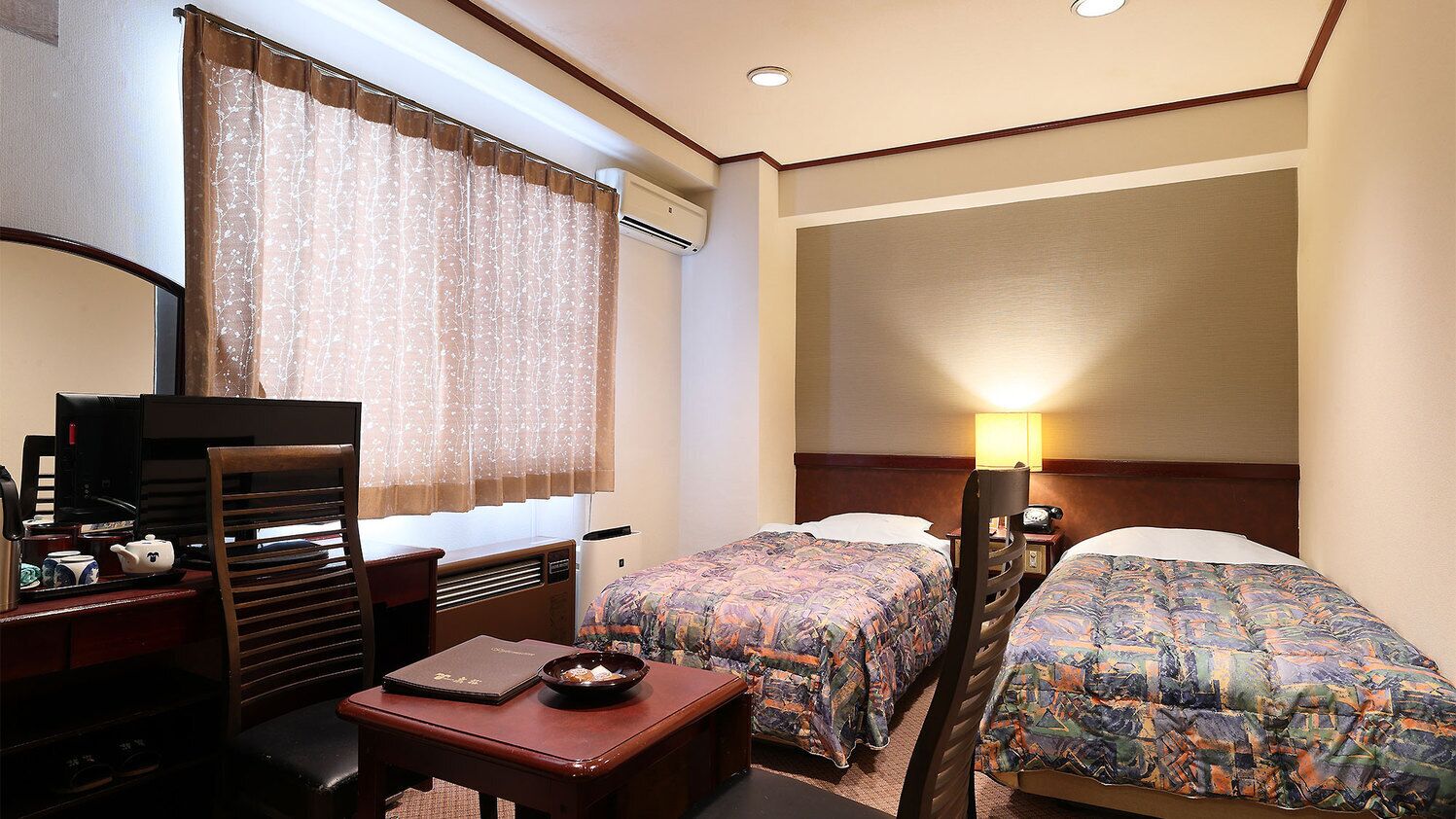 Quarto de hotel com duas camas, uma escrivaninha com cadeira, uma mesinha, uma luminária e uma janela com cortinas.