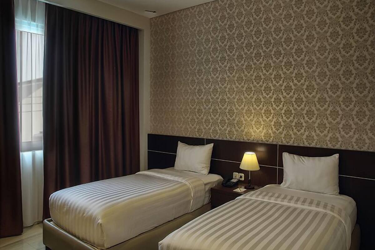 A hotel room with two beds, a patterned wallpaper, a nightstand with a lamp, and a window with curtains.