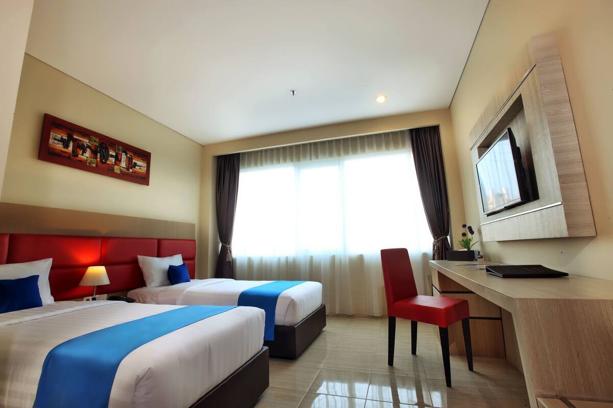 A hotel room with two beds, a desk, a TV, and a window with curtains.