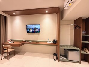 A modern hotel room with a flat-screen TV mounted on the wall, a wooden desk with a chair, a small trash bin, and a shelf with shoes and a handbag. - Hotel Asoka Luxury (Bandar Lampung)