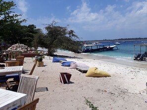 A beachside area with a table and chairs, a pile of coconuts, and a boat named 'Hugo' in the background. - Dream Divers (Gili Trawangan)