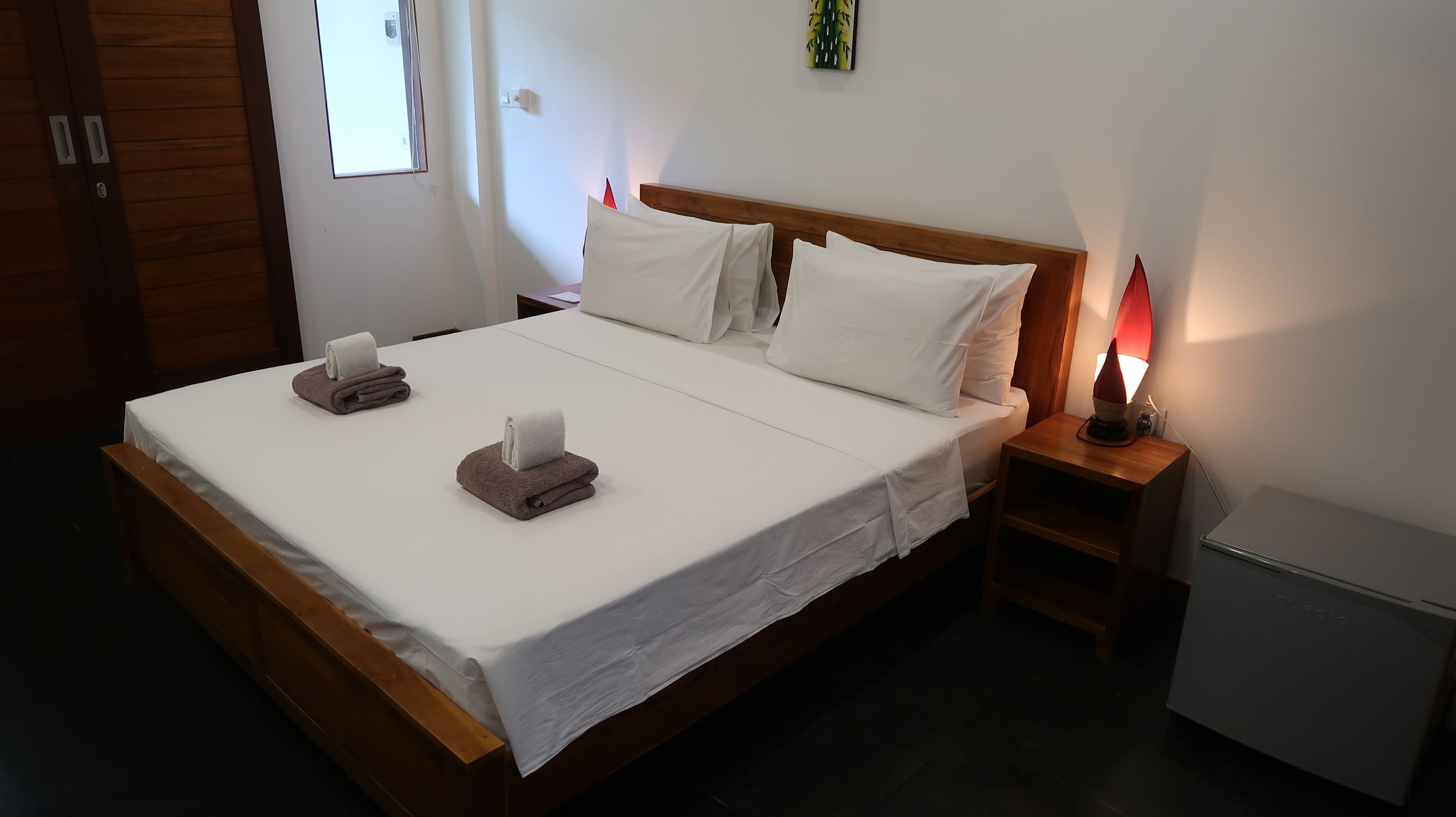Una cama bien tendida con ropa de cama blanca y marrón, una mesita de noche con lámpara y un mueble pequeño.