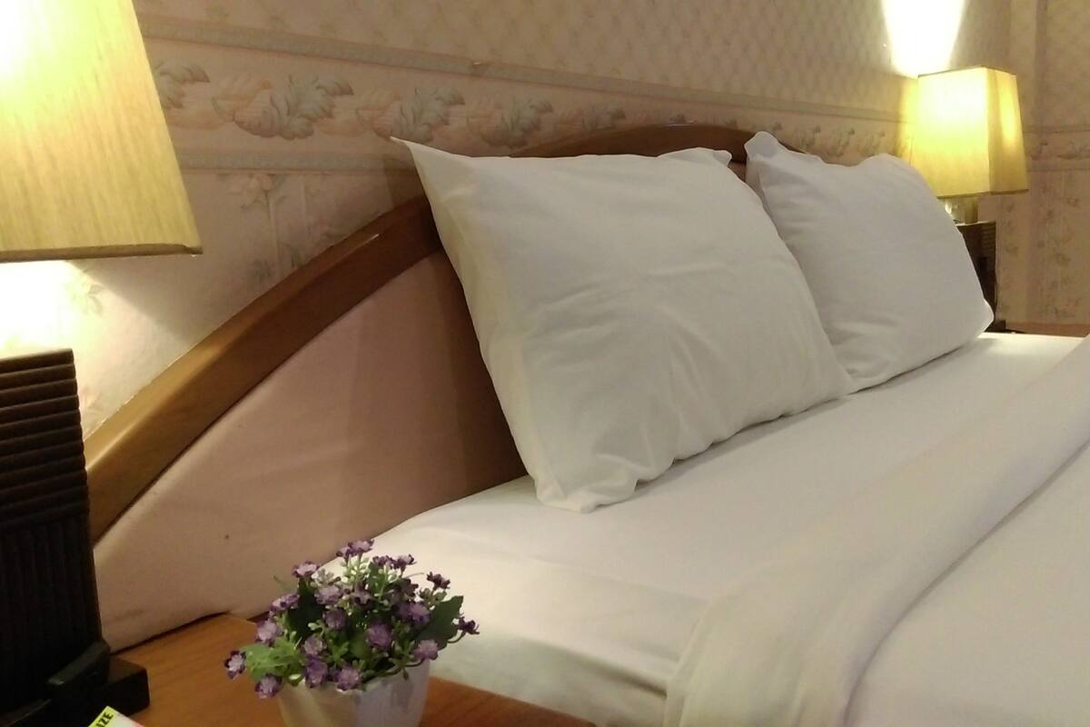 A neatly made bed with white linens, two pillows, and a small vase with purple flowers on the nightstand.