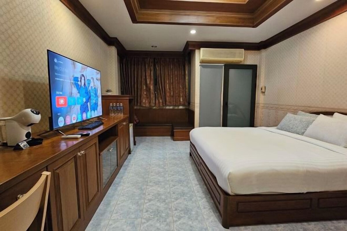 A hotel room with a large flat-screen TV on a wooden desk, a bed with white linens, and a small seating area with a chair and a table.