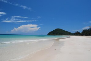 Una playa con agua turquesa cristalina, una orilla arenosa y una pequeña colina con vegetación al fondo.