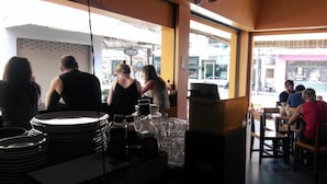En restauranginteriör med bord och stolar, en disk med staplade tallrikar och flaskor, och utsikt över gatan utanför.