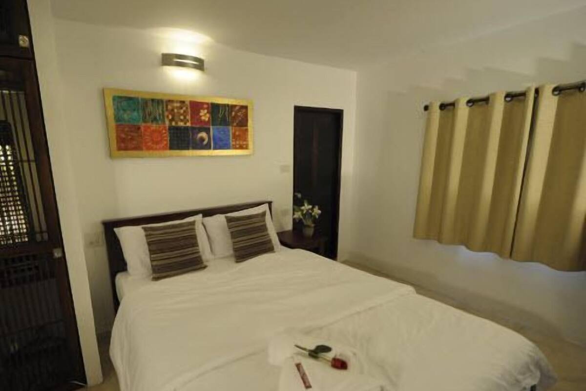 Una cama bien hecha con sábanas blancas, un cuadro abstracto de colores en la pared y una puerta cerrada con cortinas.