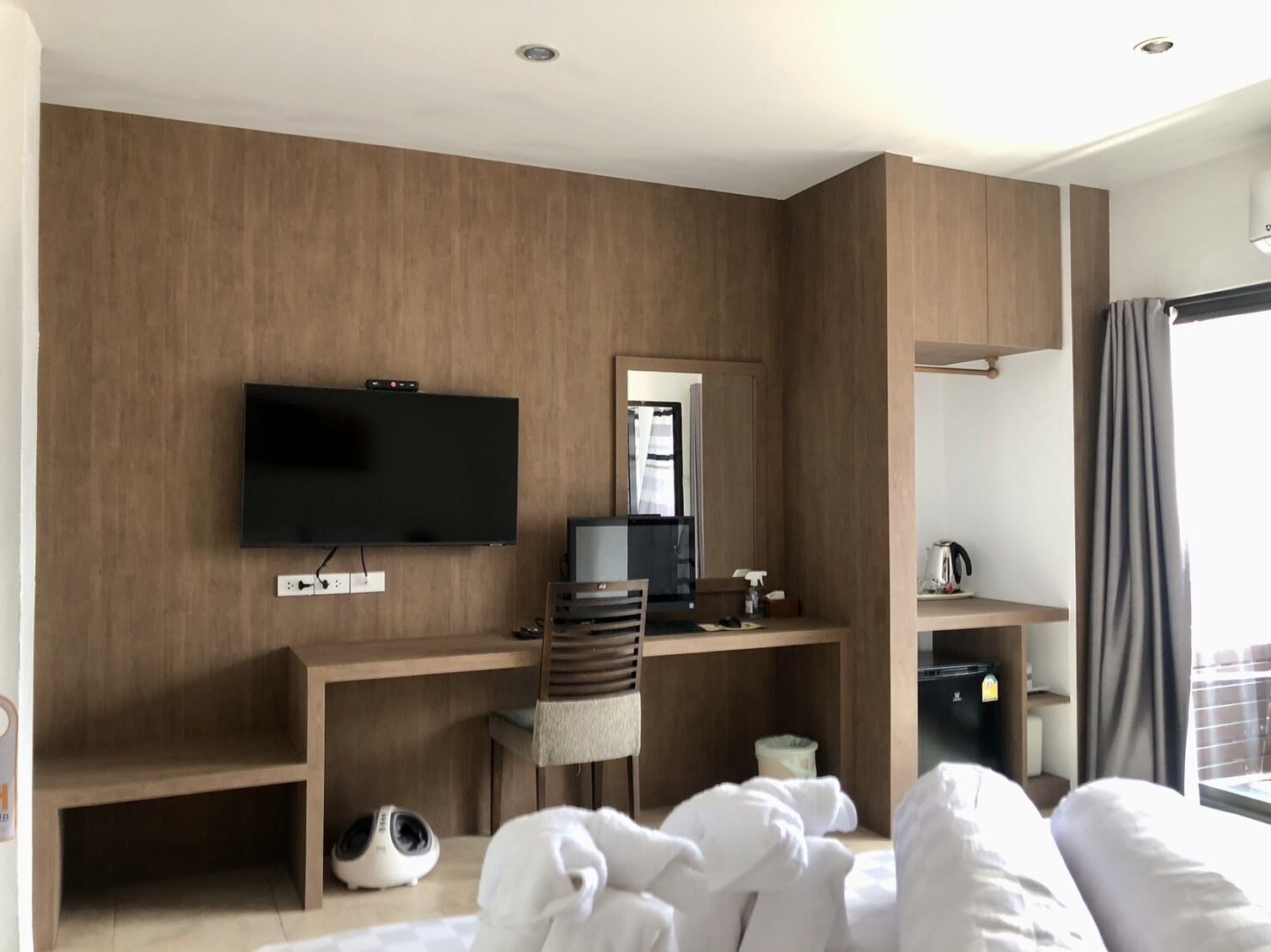 A modern hotel room with a wooden wall, a flat-screen TV, a desk with a computer, a chair, a small heater, and a large window with curtains.
