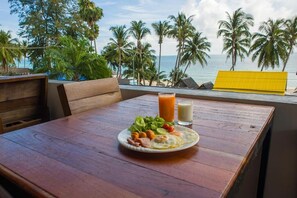 A wooden table with a plate of food and drinks, overlooking a beach with palm trees and the ocean.