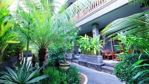 A lush garden with a pathway leading to a balcony with wooden furniture. - Letos Kubu Homestay (Kuta)