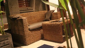 A woven sofa with a cushion, positioned in a room with a tiled floor and a window with blinds. - Letos Kubu Homestay (Kuta)