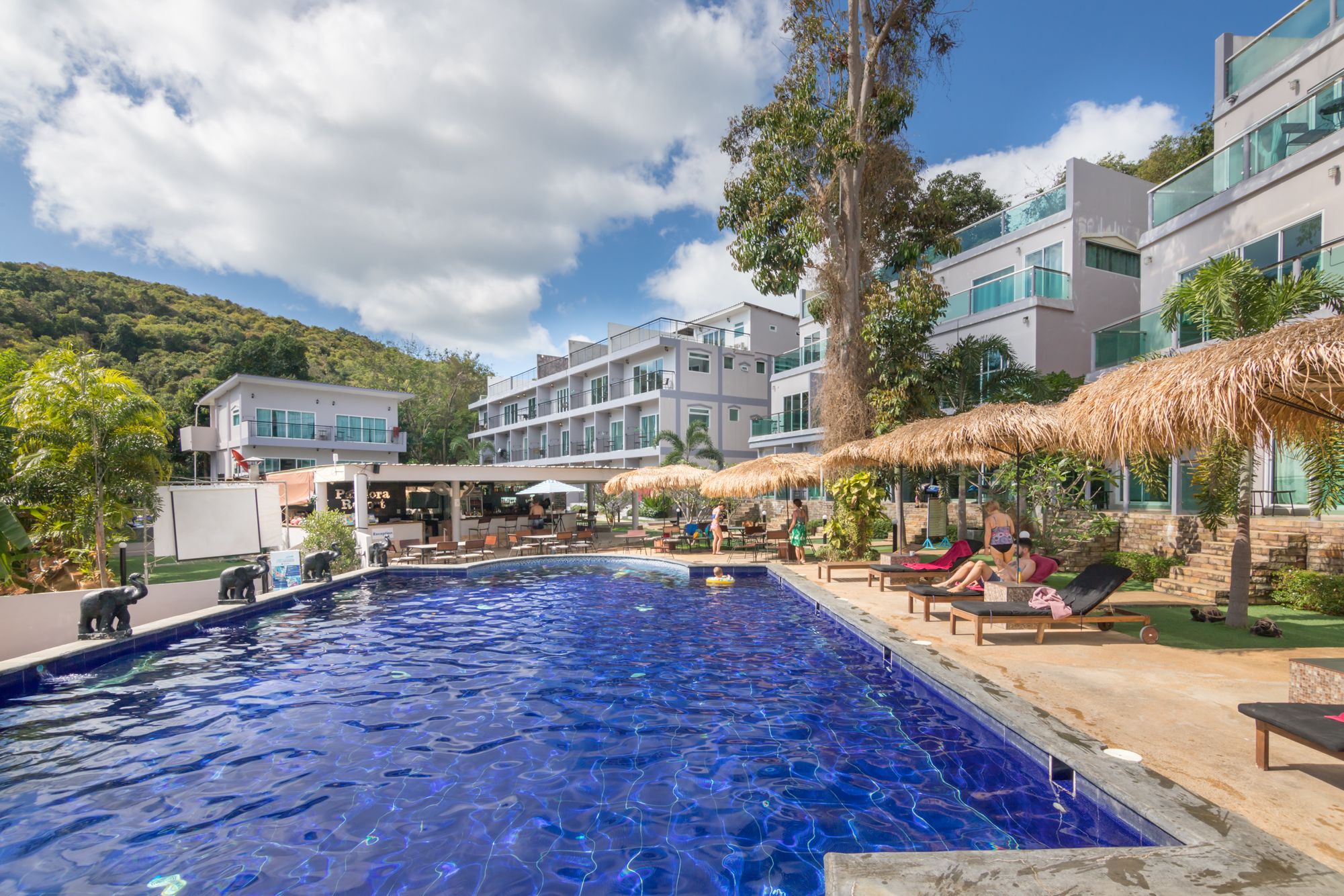 A swimming pool area with lounge chairs and umbrellas, surrounded by modern buildings and a mountainous backdrop.