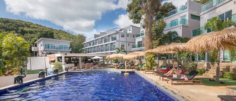 A swimming pool area with lounge chairs and umbrellas, surrounded by modern buildings and a mountainous backdrop.