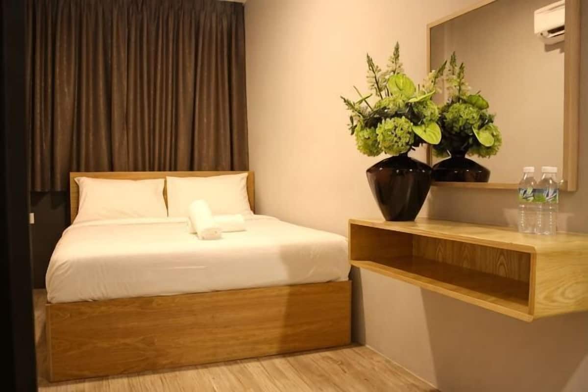 A hotel room with a bed, a wooden headboard, a bedside table with a vase of flowers, a mirror, and a shelf with bottles.