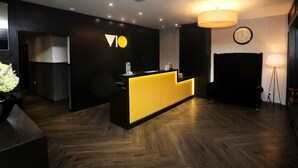 A reception area with a black and yellow desk, a clock on the wall, and a potted plant. - Vio Hotel (Kuala Lumpur)