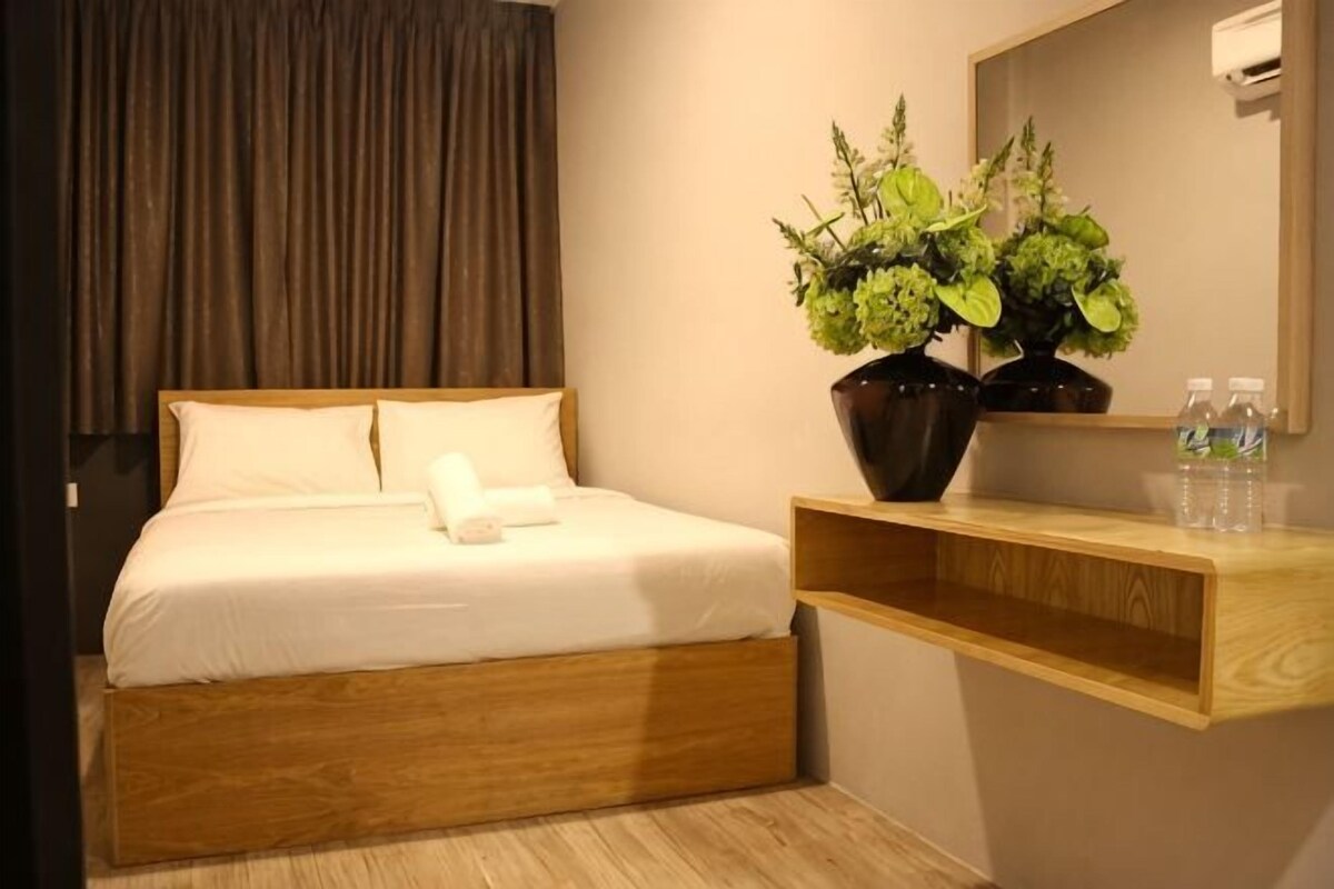 A hotel room with a bed, a wooden headboard, a bedside table with a vase of flowers, a mirror, and a shelf with bottles.