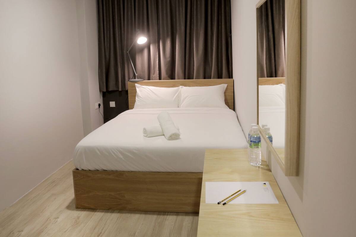 A neatly made bed with white linens, a wooden headboard, a bedside table with a bottle of water and a glass, and a mirror reflecting the room.