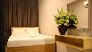 A modern hotel room with a wooden bed, a wooden nightstand, a mirror, a vase with flowers, and a shelf with water bottles. - Vio Hotel (Kuala Lumpur)