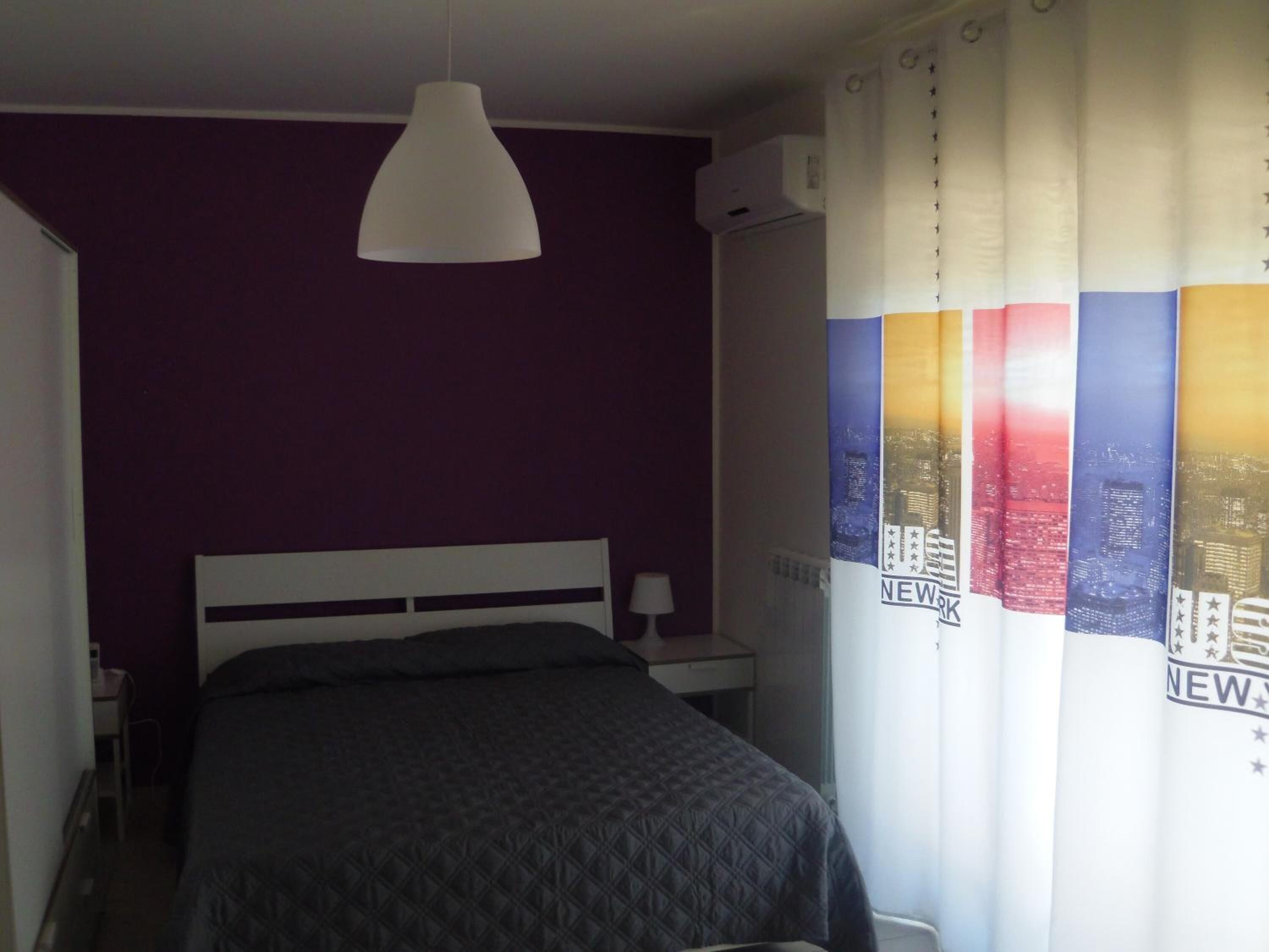A bedroom with a bed, a white pendant light, a nightstand, and a window with a curtain featuring cityscape and stars.