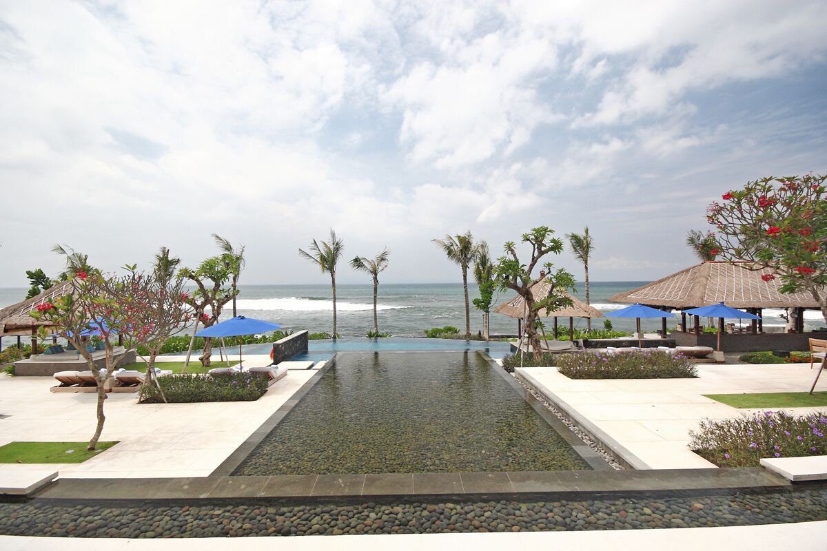 A resort pool with a clear view of the ocean, surrounded by palm trees and umbrellas.