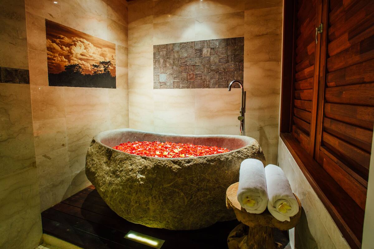 Eine Steinebadewanne, gefüllt mit roten Kieselsteinen, eine hölzerne Sitzbank mit Handtüchern und eine Wand mit einer strukturierten Mosaikverkleidung.